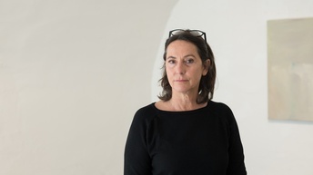Birgit Fraisl steht in einem hellen Galerieraum vor einer weißen Wand und blickt direkt in die Kamera. Sie trägt ein schwarzes Oberteil, ihre Brille liegt auf dem Kopf. Im Hintergrund ist unscharf ein großformatiges Kunstwerk zu sehen.