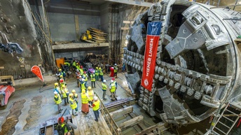 Foto einer unterirdischen Baustelle aus erhöhter Perspektive. Rechts nimmt der große, kreisförmige Schneidkopf einer Tunnelbohrmaschine einen wesentlichen Teil des Bildes ein. An der Maschine hängt ein rot-blaues Banner mit der Aufschrift „WIENER LINIEN“. Links neben der Maschine steht eine Gruppe von Personen mit Schutzhelmen und Warnwesten