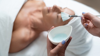 Nahaufnahme von Händen einer Person in einem Kosmetikstudio, die gerade eine Gesichtsbehandlung vorbereitet und dazu eine Creme mit Pinsel in der Hand hält