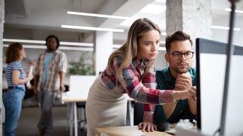 Eine Person mit langen hellbraunen Haaren und eine Person mit kurzen braunen Haaren, Bart und Brille blicken gemeinsam auf einen Monitor bei einem Schreibtisch, im Hintergrund zeigen sich weitere Personen in einem Büro