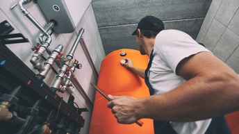 Person in Arbeitskleidung hält ein Tablet in der Hand und kontrolliert eine Messanzeige bei einem Wasserboiler in einem Heizsystem