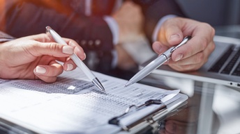 Nahaufnahme von zwei Personen und deren Händen. Sie halten beide einen Kugelschreiber in den Händen, mit dem sie ein Dokument ausfüllen, die in ein Clipboard geklemmt sind. Das Klemmbrett liegt auf einem Glastisch, auf dem auch ein Laptop ist