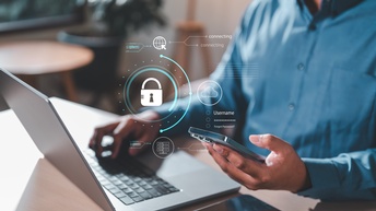 Person sitzt vor einem Laptop bei einem Schreibtisch und hält ein Handy in der linken Hand. Über Handy und Laptop schweben verschiedene Symbole wie ein Schloss in einem Kreis oder eine Weltkugel mit einem Pfeil