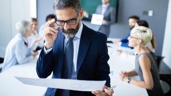 Person in Businesskleidung mit Brille blickt auf ein Dokument in seinen Händen, im Hintergrund sitzen weitere Personen an einem Tisch und eine weitere Person moderiert das Gespräch