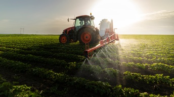 Ein roter Traktor mit breitem schwarzen Aufsatz mit Spritzdrüsen, der über Feld mit grünen Pflänzchen fährt und Flüssigkeit darauf sprüht.