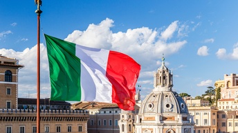 Die Flagge Italiens im Wind wehend unter blauem Himmel mit Wolken, im Hintergrund historische Gebäude.
