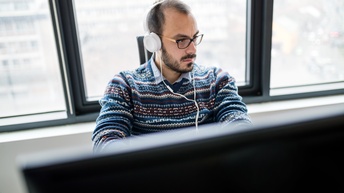 Person mit Kopfhörer und Brille sitzt an einem Computerarbeitsplatz, im Hintergrund befindet sich eine Fensterfront