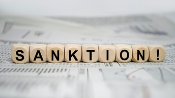 Neun Würfel liegen nebeneinander auf bedrucktem Papier. Auf jedem Würfel ist ein Buchstabe, wodurch sich das Wort Sanktion ergibt. Am letzten Würfel ist ein Rufzeichen