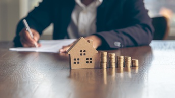 Auf einem Tisch steht ein kleines Modellhaus. Daneben stehen nebeneinander fünf, unterschiedlich hohe Münzstapel. An dem Tisch sitzt eine Person, vor der ein Zettel ist, auf dem die Person etwas notiert