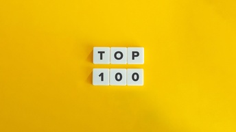 Illustration eines gelben Hintergrund. In der Mitte liegen in zwei Reihen jeweils drei weiße Würfel. In der oberen Reihe ist auf jedem Würfel ein Buchstabe, wodurch sich das Wort Top ergibt. In der unteren Reihe sind Zahlen, wodurch sich 100 ergibt