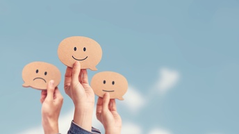 Vor blauem Himmel sind drei Hände in die Luft gestreckt. Jede Hand hält ein Smiley. Ein Smiley lächelt, eines blickt neutral und das dritte zieht die Mundwinkel nach unten