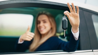 Nahaufnahme von einer einem Autoschlüssel, der von einer Hand gehalten wird, in der Unschärfe sitzt eine Person in einem Auto und zeigt mit dem Daumen nach oben