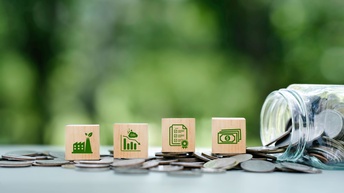 Holzwürfel mit Symbolen für Industrie, CO₂-Reduktion, Zertifizierung und finanzielle Förderung stehen auf verstreuten Münzen. Rechts liegt ein umgekipptes Glas mit herausgerollten Münzen. Der unscharfe grüne Hintergrund vermittelt einen Bezug zu Nachhaltigkeit, Umwelt- und Klimaschutzmaßnahmen in Unternehmen.