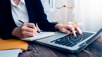 Topshot der Hände einer Person, die mit einem Kugelschreiber in ein Notizbuch schreibt. Vor ihr ist ein Laptop auf einem braunen Holztisch aufgeklappt. Man sieht grafische Symbole von  Personen, Notizen und einem Parlament. Neben ihr  steht eine Waage.