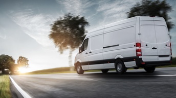 Ein weißer Kleintransporter fährt auf der rechten Spur einer asphaltierten Straße. Neben der Straße sind grüne Wiesen sowie grüne Bäumen
