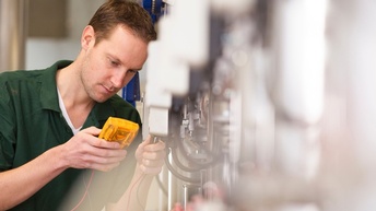 Person mit kurzen Haaren und grünem Poloshirt misst mit einem Gerät einen Wert einer industriellen Anlage