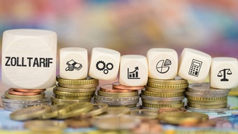 Nahaufnahme von fünf Münzstapeln nebeneinander. Auf den Münzen liegen sieben weiße Würfel. Auf dem ersten steht Zolltarif. Auf den anderen sind Symbole wie ein Taschenrechner oder eine Waage