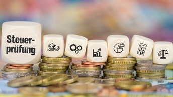 Weiße Würfel mit schwarzem Schriftzug Steuerprüfung und Symbolen zum Thema Steuer wie Geldscheine, Diagramme und Taschenrechner platziert auf Euro-Münzstapeln