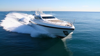 Weiße Yacht fährt über Wasser unter blauem Himmel, weiße Gischt entlang der befahrene Strecke des Bootes und am Bug, im Hintergrund Inselgruppe