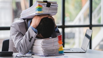 Eine Person in einem hellgrauen Sakko sitzt an einem Tisch und hat ihren Kopf auf einem hohen Stapel Papier abgelegt. Auf ihrem Kopf liegt ein weiterer Stapel Papiere. Neben ihr steht ein aufgeklappter Laptop