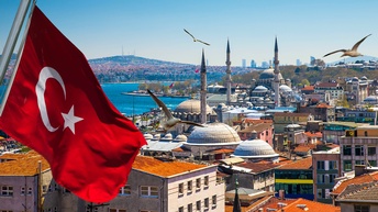 Im Bildvordergrund ist in der linken Hälfte eine Flagge des Landes Türkei an einer Metallstange. Im Hintergrund sind die Dächer verschiedener Gebäude sowie einer Moschee mit drei Türmen. Über der Stadt fliegen Möwen.