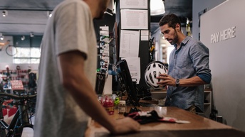 Person steht hinter Verkaufstheke und blickt auf Computer, hält weißen Sporthelm in Händen, im Vordergrund verschwommen weitere Person, ringsum Ausschnitte von Fahrrädern