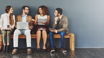 Vier Personen sitzen auf einer Holzbank in einem Raum. Sie blicken sich gegenseitig an und interagieren miteinander. Auf ihren Schößen liegen von links nach rechts: Papiere, ein Laptop, ein Notizblock und ein Smartphone, ein Tablet