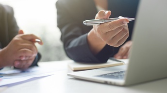 Ausschnitt der Hände zweier Personen, die an einem Tisch sitzen. Beide halten jeweils einen Stift. Vor ihnen auf dem Tisch steht ein aufgeklappter Laptop