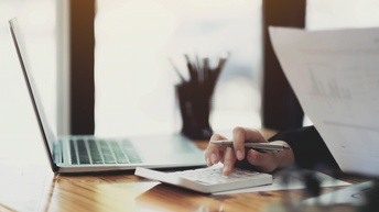 Nahaufnahme der rechten Hand einer Person, die an einem Schreibtisch sitzt und auf einem Taschenrechner tippt. In der linken Hand hält sie einen Zettel mit Statistiken. Neben dem Taschenrechner ist ein aufgeklappter Laptop