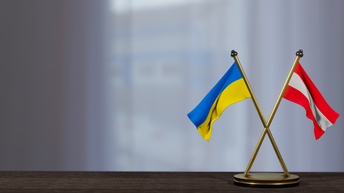 In der rechten Bildhälfte sind zwei sich kreuzende, vergoldete Metallstäbe, die sich kreuzen und unten an einer Scheibe montiert sind. Auf einem Stab ist die ukrainische Flagge, auf dem anderen die österreichische