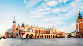 Auf einem gepflasterten Platz ist ein längliches, zweistöckiges, historisches Gebäude. Links daneben ist eine Kirche. Rechts daneben ist ein Turm