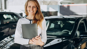 Lächelnde Person mit Mappe in Händen lehnt an dunklem Auto