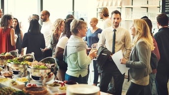 Mehrere Personen in Bürokleidung stehen in einem Raum in kleinen Gruppen und unteragieren miteinander. In der linken Bildhälfte ist ein gedeckter Tisch mit Gefäßen, befüllt mit mehreren Speisen. Im Hintergrund hängt eine dunkle, beschriftete Tafel