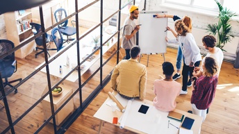 Sieben Personen stehen um eine kleine Tafel. Zwei Personen halten Stifte. Die anderen blicken auf die Tafel