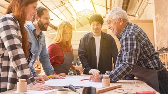 Hier zu sehen ist eine 5-köpfige Gruppe an Tischlern. Ein älterer Herr ist der Meister, welcher den anderen etwas auf einem Plan erklärt. Auf dem Tisch um den sie stehen, findet man Holz und Werkzeug. Im Hintergrund sieht man die Werkstatt.
