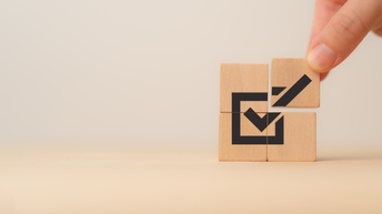 Vier Holzwürfel liegen auf einem Holztisch und  bilden ein Quadrat. Auf jedem Würfel ist ein Teil einer Abbildung, sodass sich ein Haken in einem Quadrat ergibt, Ein Würfeln wird mit zwei Fingern leicht angehoben.