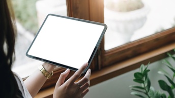 Detailansicht eines Tablets mit weißem Display, das von einer Person vor Fenster in Raum gehalten wird, im Ausschnitt verschwommen Zimmerpflanze