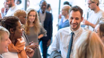 Im Vordergrund stehen fünf Personen in einem Kreis und unterhalten sich. Im Hintergrund stehen viele weitere Personen in Kleingruppen und unterhalten sich