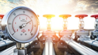 Detailansicht eines Manometers, im Hintergrund verschwommen Rohre und Justierungsräder unter wolkenverhangenem Himmel