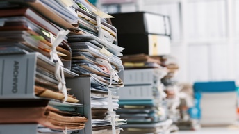 In der linken Bildhälfte sind viele Reihen übereinander gestapelter Aktenordner. In den Ordnern sind jeweils viele Zettel und Unterlagen