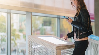 Person in Lederjacke steht vor großem Touchscreen, bedient dieses und blickt darauf, in einer Hand hält die Person ein Smartphone, im Hintergrund verschwommen Fensterfront