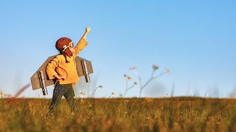 Ein Kind mit einer Fliegermütze und einer Fliegerbrille steht auf einer Wiese und streckt den linken Arm nach oben. Das Kind hat am Rücken gebastelte Flügel. Der Himmel ist blau