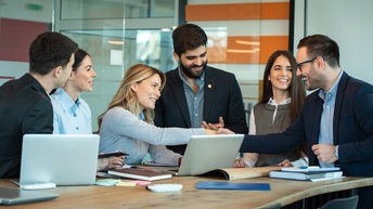 Mehrere lächelnde Person um Tisch versammelt, auf dem aufgeklappte Laptops stehen und Arbeitsutensilien liegen; zwei Personen reichen sich die Hand