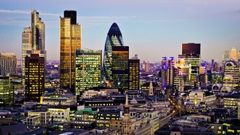 Panoramaaufnahme der Innenstadt Londons mit historischen Gebäuden und Wolkenkratzern. Die Gebäude sind teilweise beleuchtet. Dahinter erstreckt sich ein Teil der Stadt