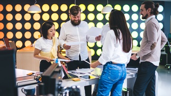 Fünf Personen stehen in einem Büro um einen Tisch. Zwei der Personen halten jeweils einen Zettel. Auf dem Tisch liegen ein Tablet, Post-its, ein Laptop sowie andere Büroutensilien. Die Personen reden miteinander