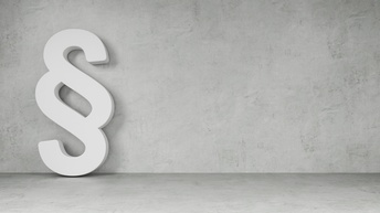 Vor einer grauen Betonwand lehnt ein weißes Paragraphen-Symbol in der linken Bildhälfte. Das Symbol ist dreidimensional.