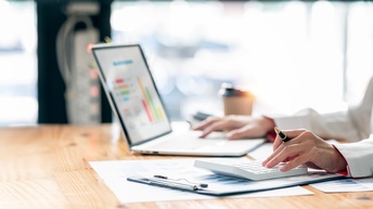 Seitliche Aufnahme einer Person, die an einem Tisch sitzt. Mit der linken Hand tippt sie auf einem Taschenrechner. Mit der rechten Hand bedient sie einen aufgeklappten Laptop, auf dem verschiedene Diagramme sind