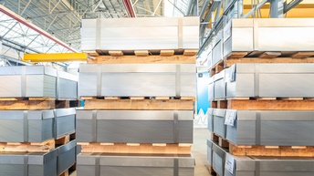 In einer hohen Halle stehen im Vordergrund drei Stapel nebeneinander. Bei jedem Stapel sind rechteckige Metallböcke, die durch helle Paletten voneinander getrennt sind. Pro Stapel sind drei oder vier Metallblöcke übereinander gestapelt