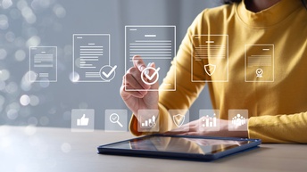 berkörper einer Person in einem gelben Oberteil an einem Tisch mit einem Tablet. Die Person berührt mit einem Finger eine eingeblendete grafische Benutzeroberfläche. Darüber sind mehrere stilisierte Dokumente mit Symbolen wie Häkchen