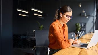 Seitliche Aufnahme einer Person mit oranger Bluse, Brille und Kopfhörern, die an einem langen Tisch neben einem Fenster sitzt und auf den Bildschirm eines Laptops blickt
